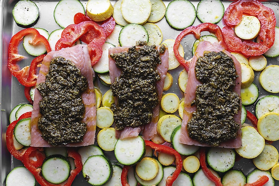 raw salmon filets covered with pesto on a sheet pan full of veggies