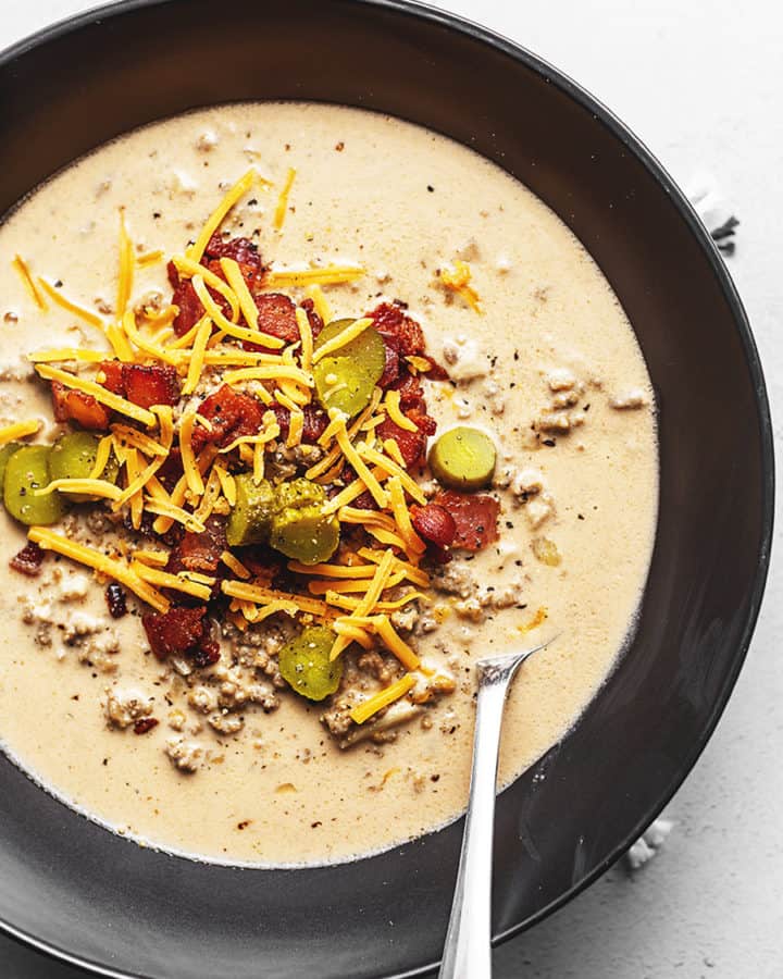 A CREAMY SOUP WITH HAMBURGER, BACON, CHEESE, AND PICKLES IN A BLACK BOWL