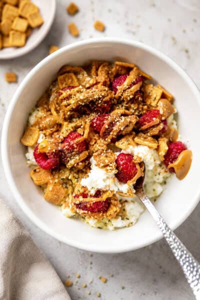 Healthy cottage cheese breakfast bowl topped with raspberries, crunchy cereal, almond butter, and hemp hearts in a white bowl.