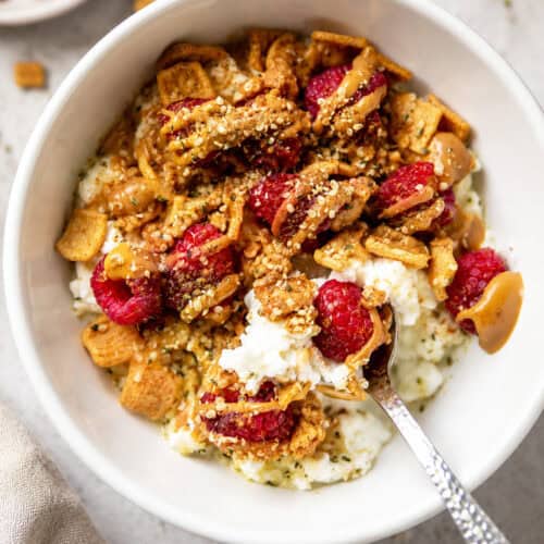 Healthy cottage cheese breakfast bowl topped with raspberries, crunchy cereal, almond butter, and hemp hearts in a white bowl.
