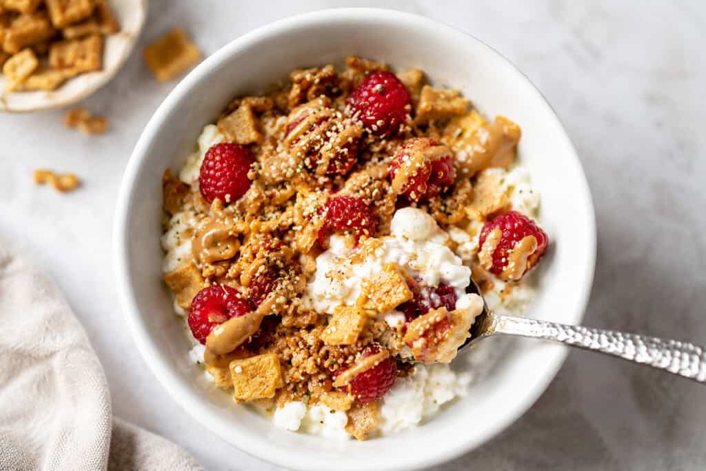 Healthy cottage cheese breakfast bowl with raspberries, crunchy cereal, almond butter, hemp hearts, and cinnamon in a white bowl.