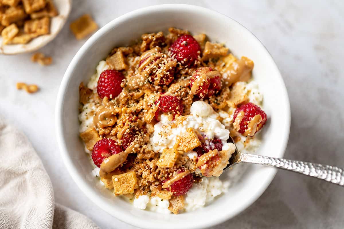 Healthy cottage cheese breakfast bowl with raspberries, crunchy cereal, almond butter, hemp hearts, and cinnamon in a white bowl.