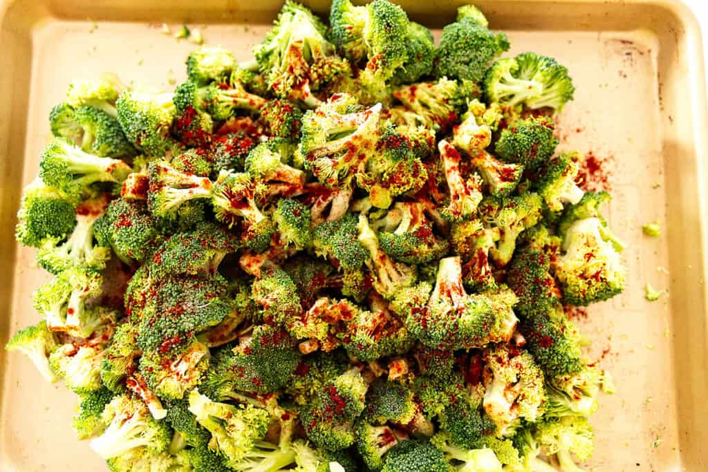 fresh broccoli, seasonings and oil on a sheet tray ready to be roasted in the oven